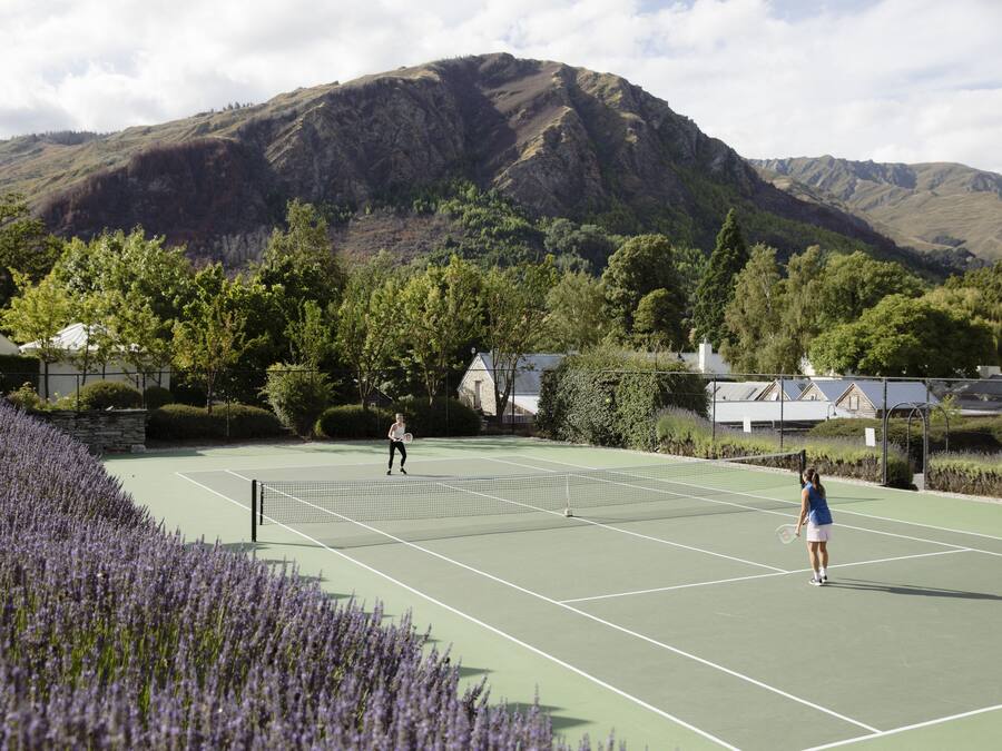 Ein Tennisplatz mit Bergelandschaft im Hintergrund, umgeben von Bäumen und einer Steinmauer.