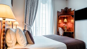 Hotellihuone, jossa on sänky, kaksi yöpöytävalaisinta, puinen yöpöytä, peili ja ikkuna, jossa on läpikuultavat verhot.