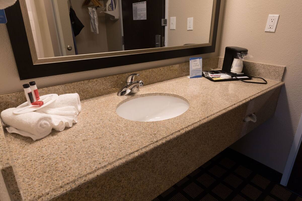 Et badeværelse på et hotel med bordplade i granit, en hvid vask, et spejl og toiletartikler.