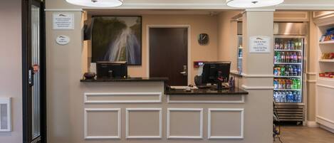 A Baymont Inn & Suites reception area with a check-in counter, a refrigerator stocked with beverages, and a clock on the wall.