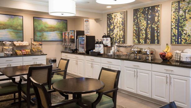 A modern kitchen with white cabinets, a black countertop, and a dining area with wooden chairs. There are paintings on the walls, a coffee machine, and a fruit bowl on the counter. - Baymont by Wyndham Detroit Airport/Romulus (Romulus)