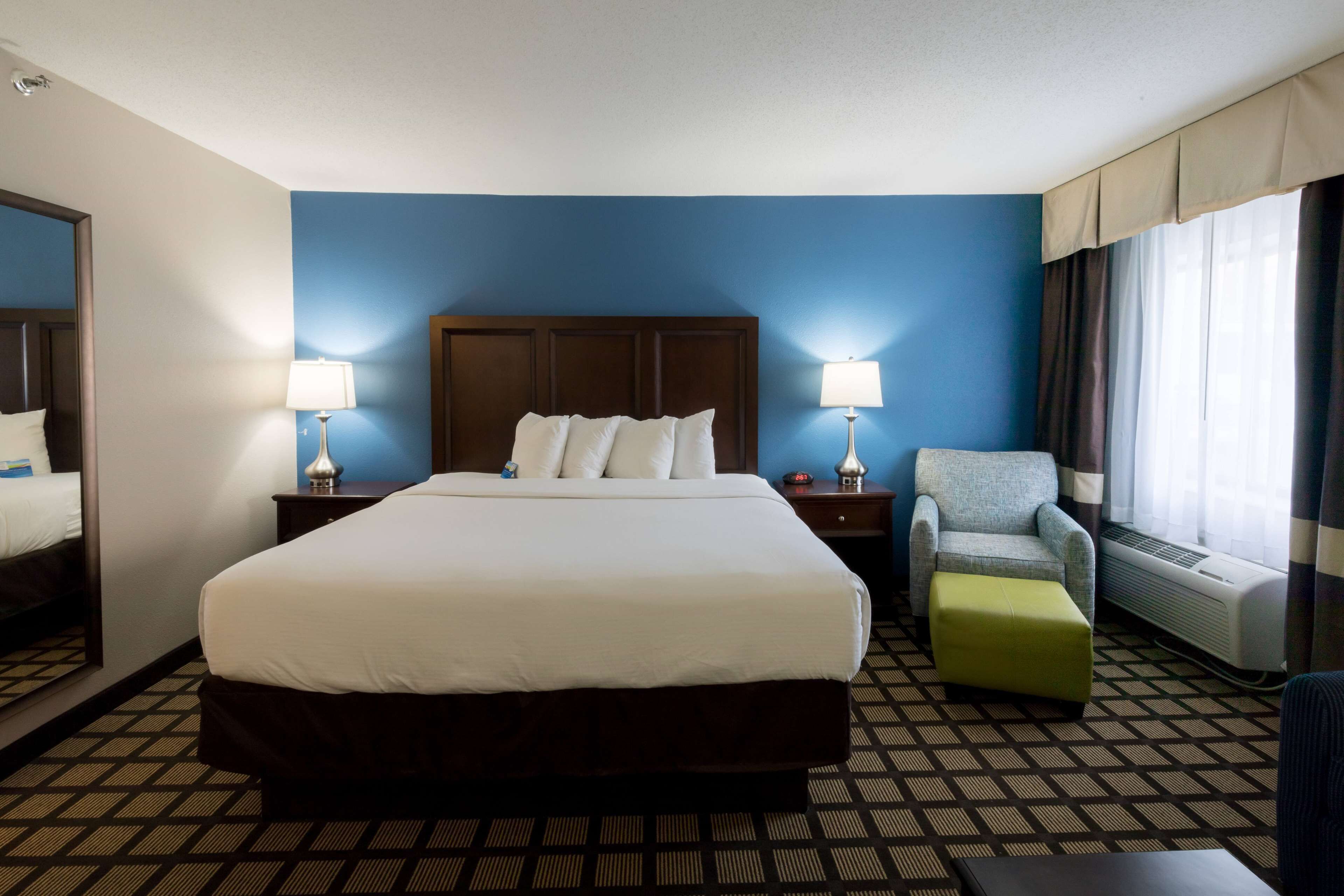 Een hotelkamer met een groot bed, twee nachtkastjes met lampen, een stoel, een klein voetenbankje en een raam met gordijnen.