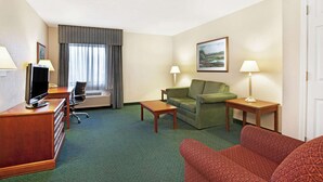 Une chambre d’hôtel avec une moquette verte, un bureau en bois avec une télévision, un canapé vert, un canapé rouge à motifs, deux tables de chevet avec des lampes et une fenêtre avec des rideaux.