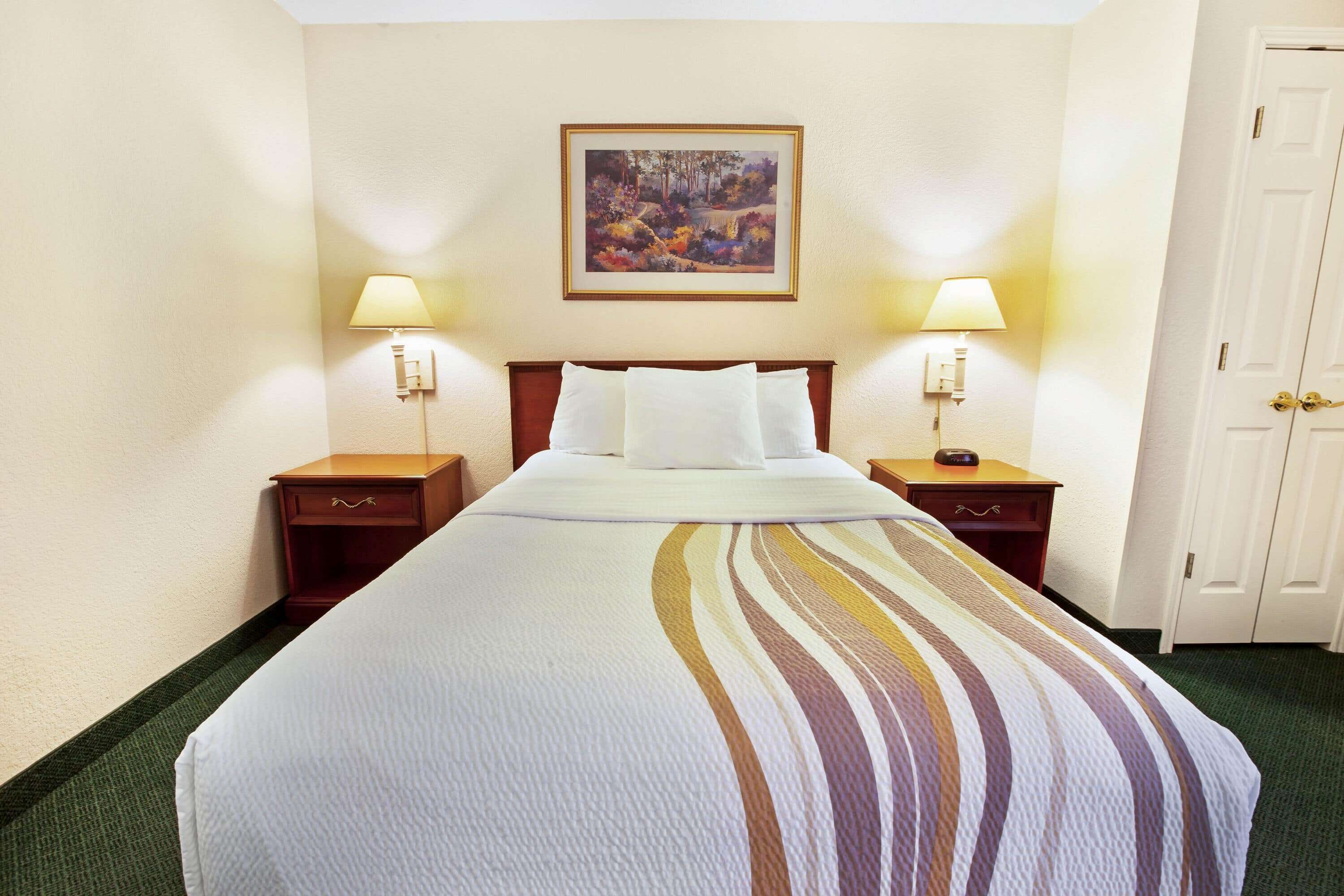 A hotel room with a neatly made bed, two bedside tables with lamps, a painting on the wall, and a door to the right.