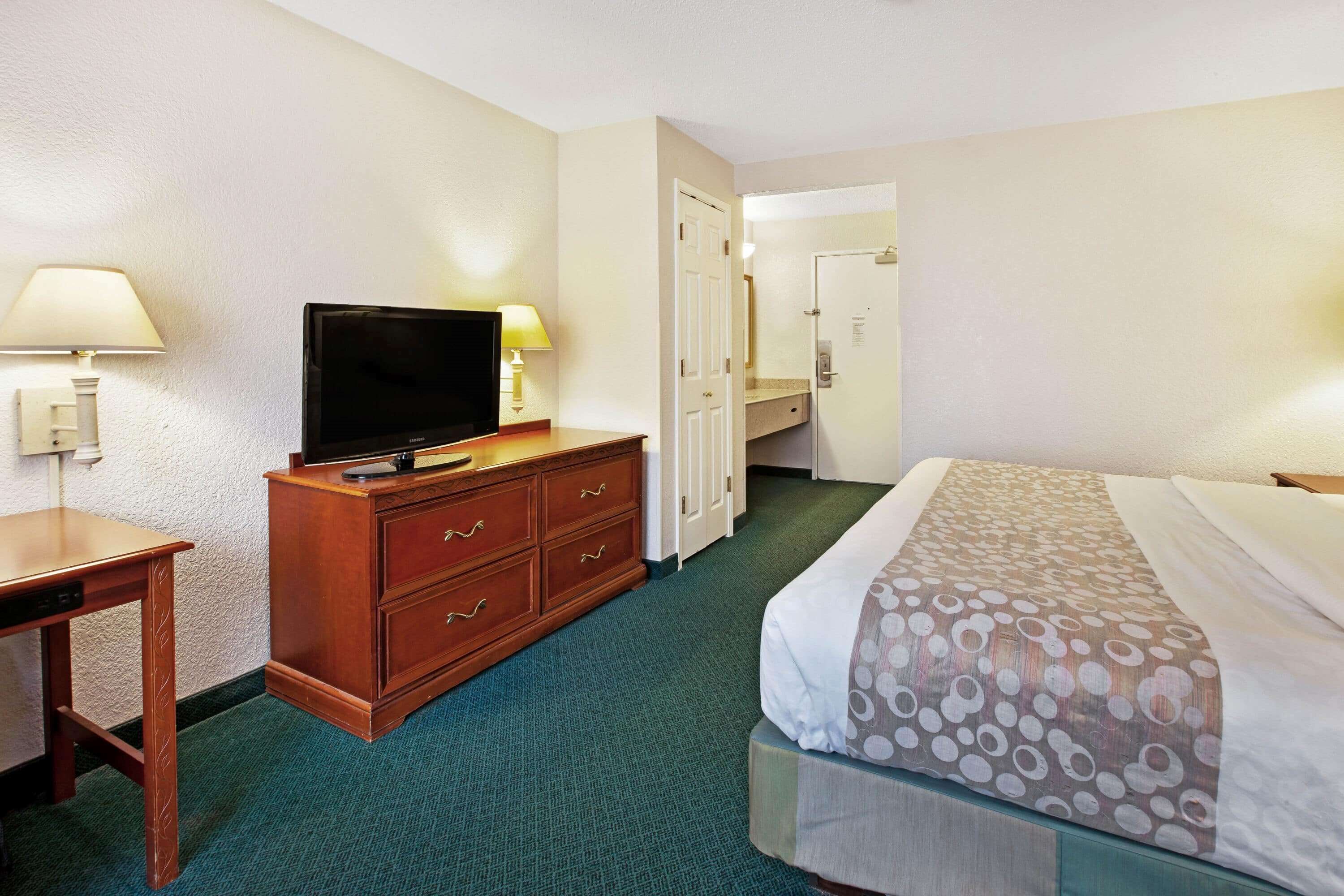 A hotel room with a bed, a TV on a wooden dresser, a desk, and a door leading to a bathroom.