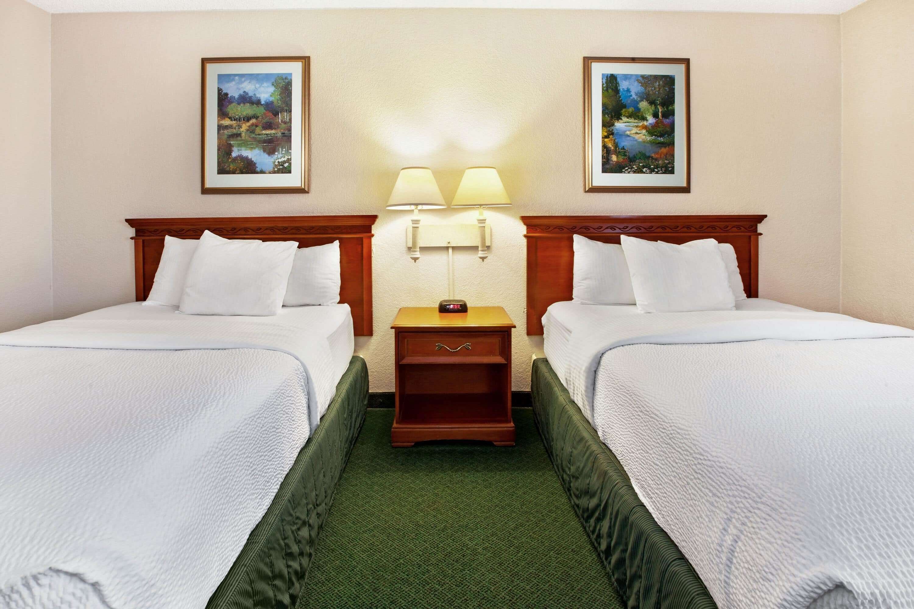 A hotel room with two single beds, a nightstand with lamps, and two framed landscape paintings on the walls.