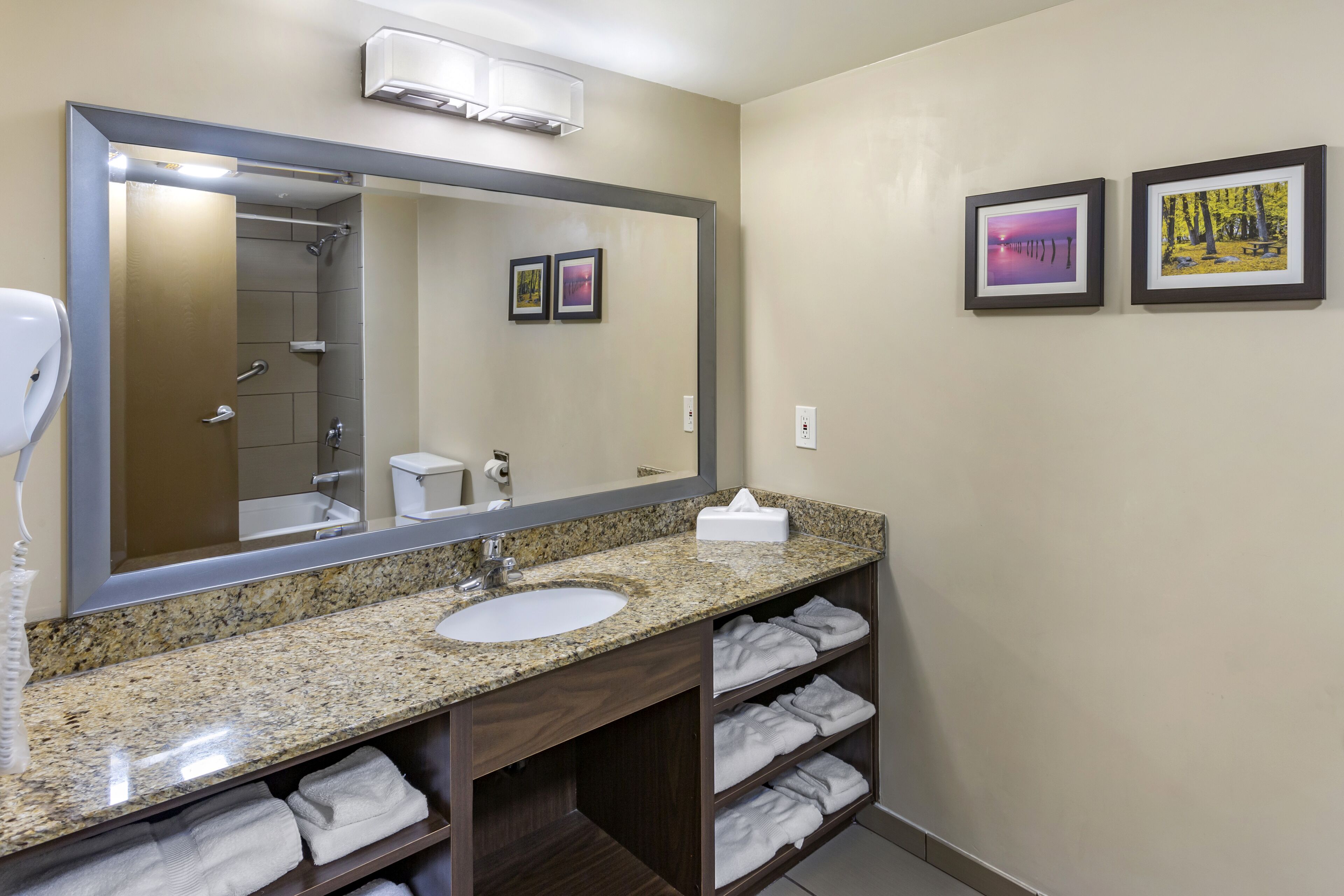 Une salle de bain avec un lavabo doté d’un plan de travail en granit, un grand miroir, un porte-serviettes et deux tableaux encadrés accrochés au mur.