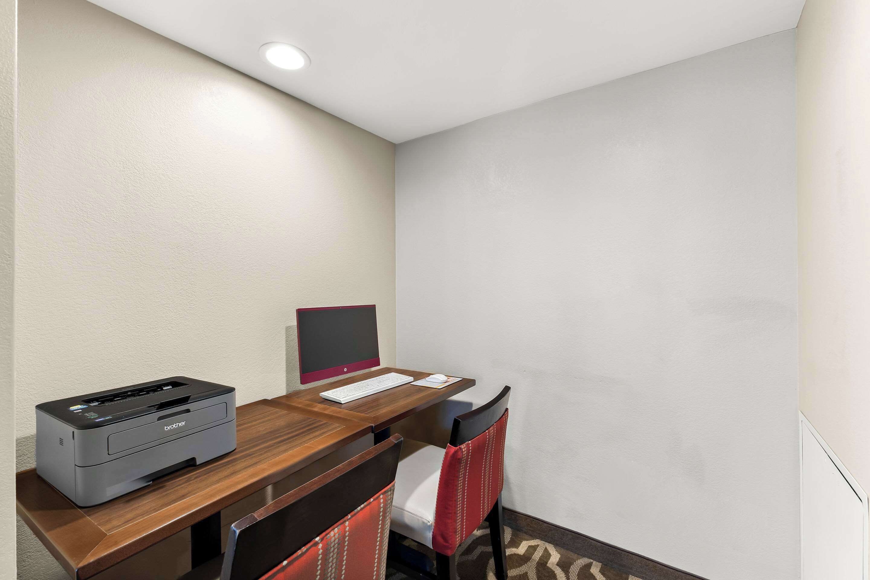 A hotel room with a wooden desk, a Brother printer, a computer monitor, and a red chair.