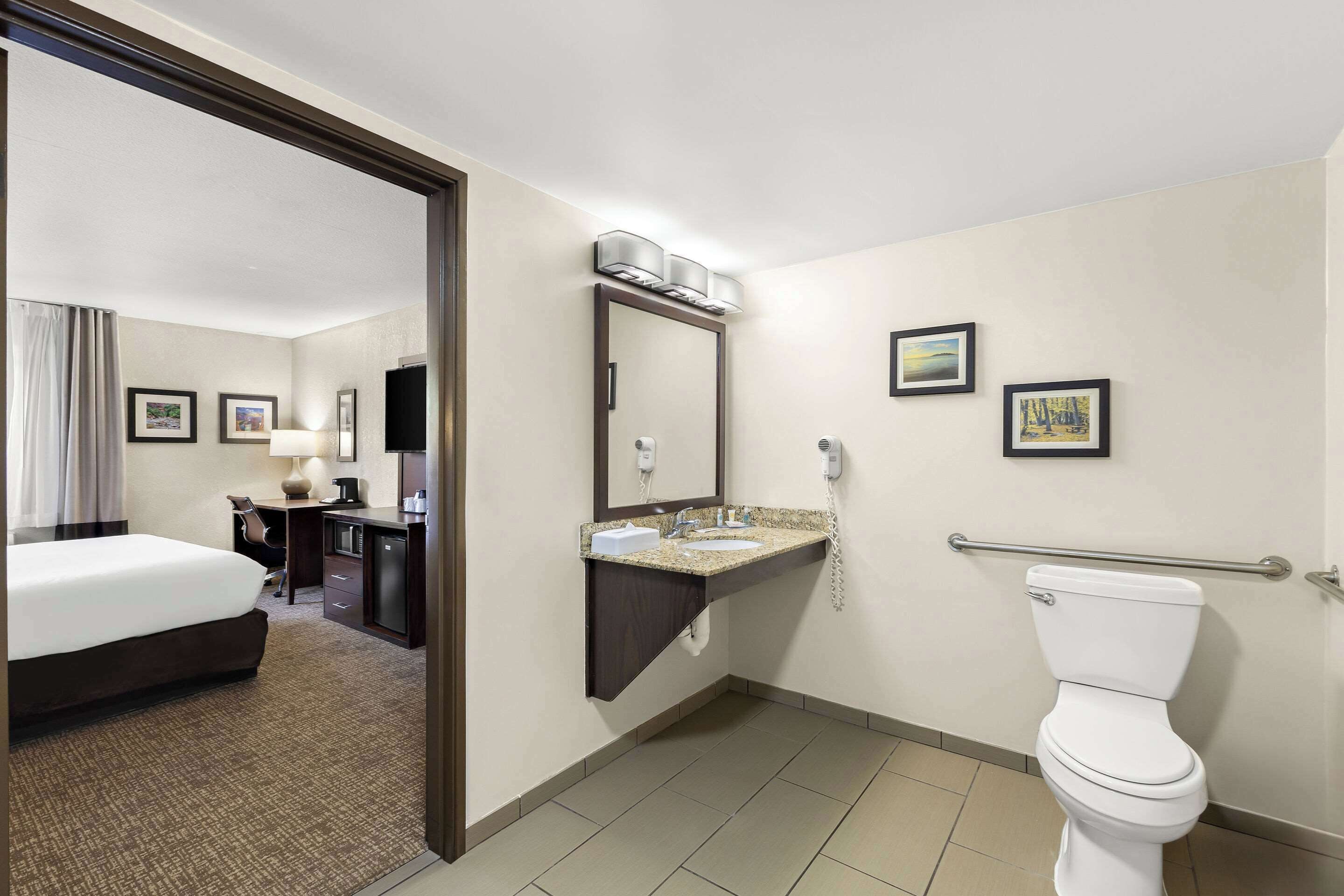 A hotel bathroom with a toilet, sink, and mirror. There is a bed and desk in the adjacent room.