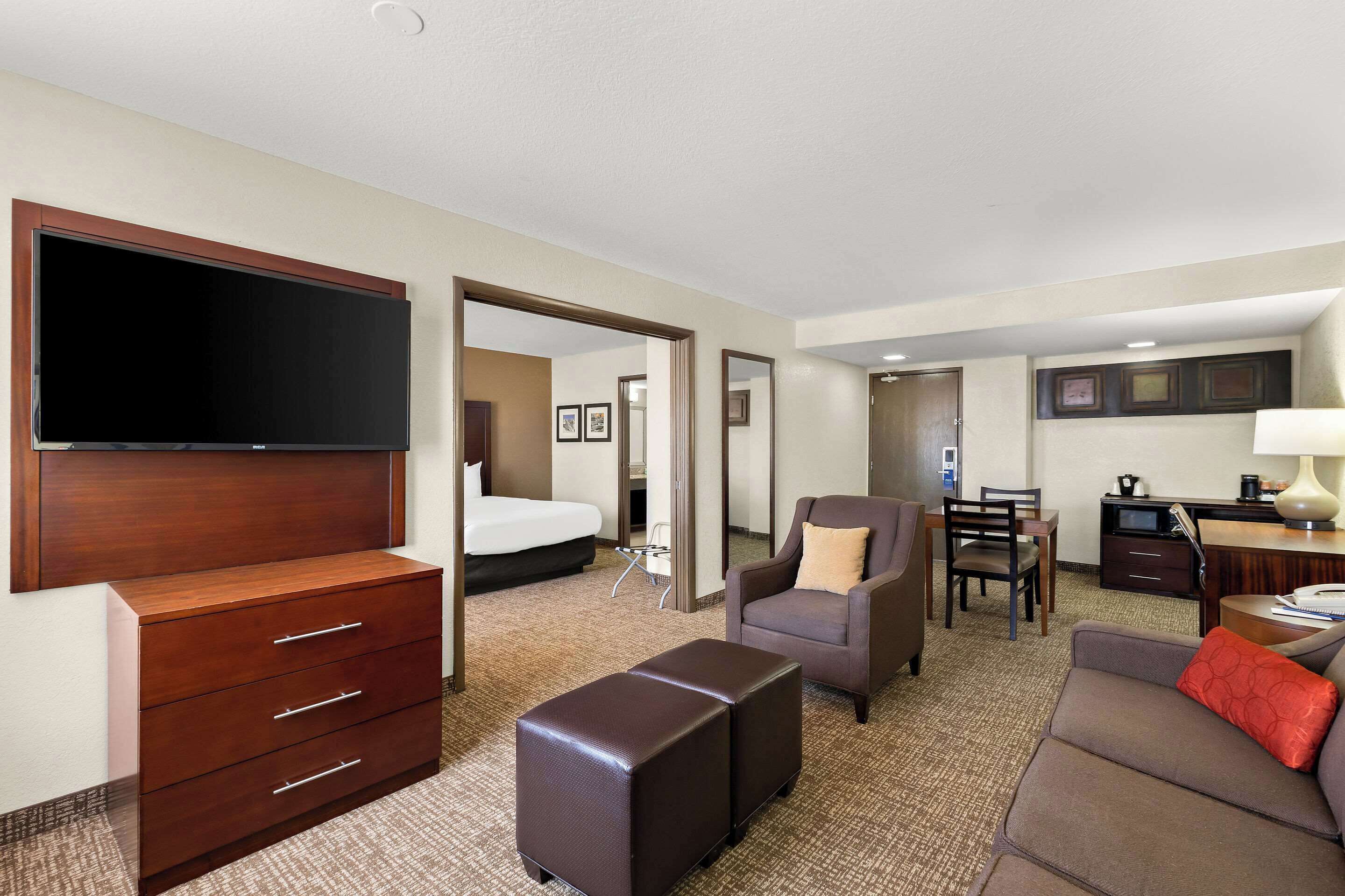 A hotel room with a flat-screen TV, a wooden dresser, a sofa, a chair, a small table, and a desk with a lamp.