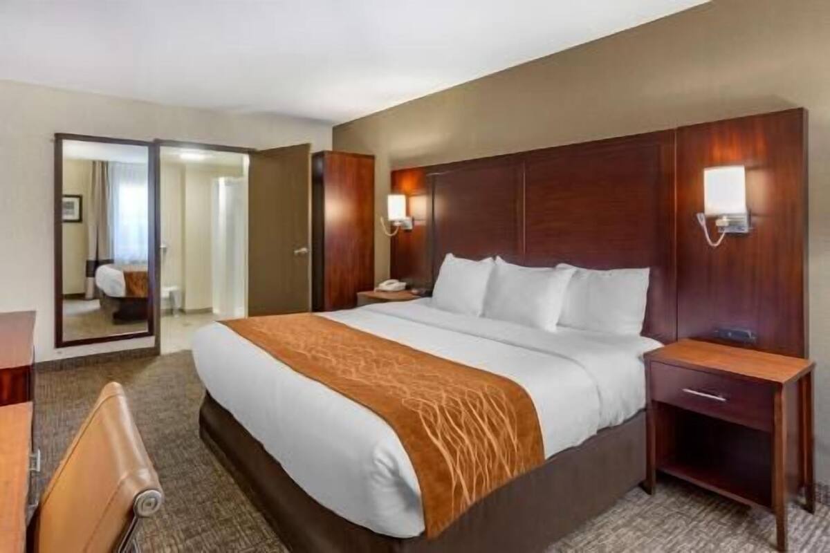Une chambre d’hôtel avec un grand lit, une tête de lit en bois, des tables de chevet, un miroir et une porte donnant sur une autre pièce.