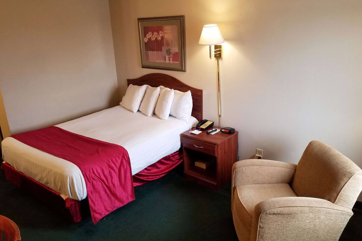 A hotel room with a bed, a nightstand with a lamp, a chair, and a framed picture on the wall.