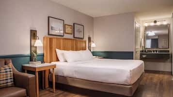 Habitación de hotel con cama, mesita de noche con lámpara, una silla, cabecera de madera y cuadros en la pared.