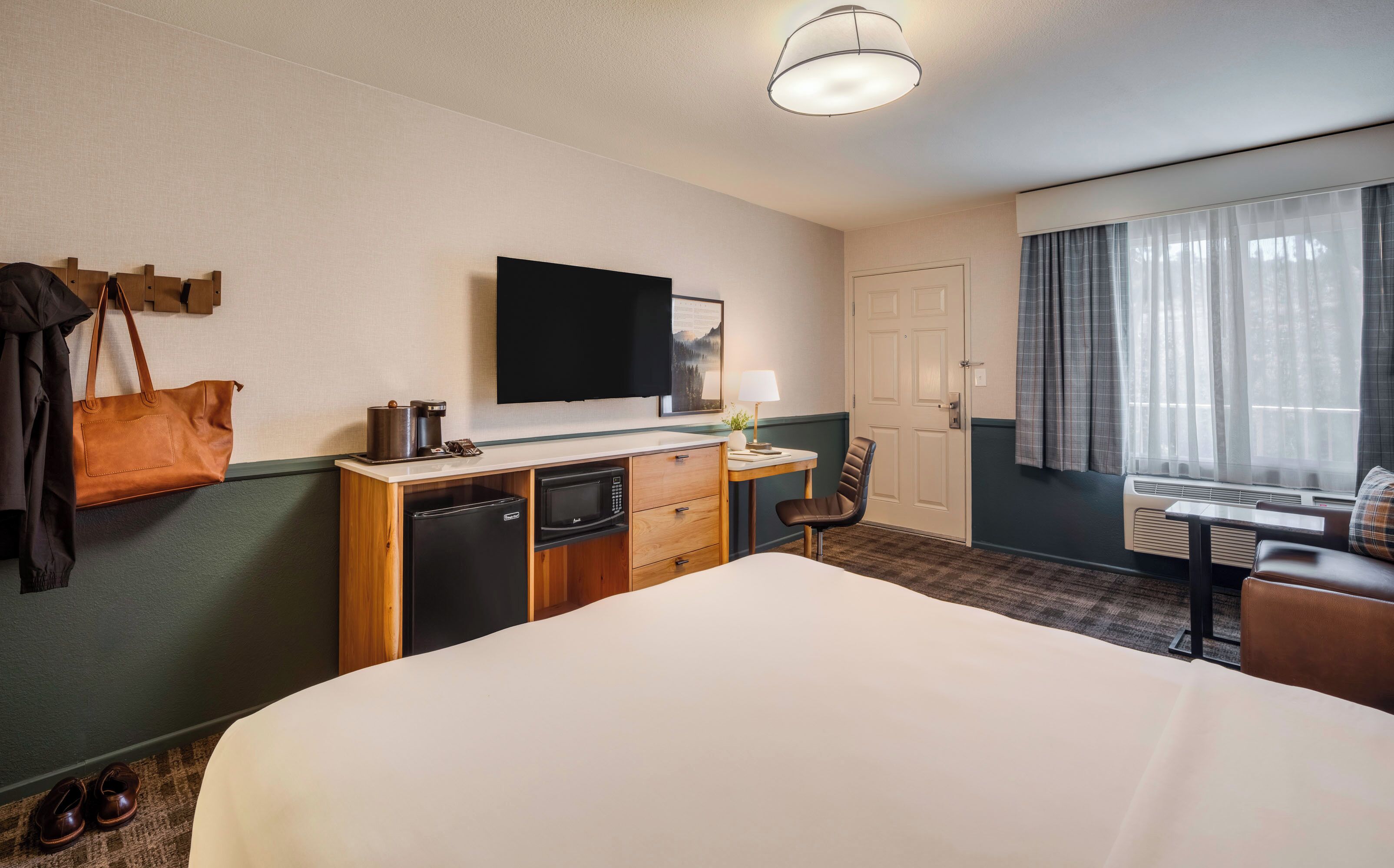Habitación de hotel con cama, escritorio con lámpara, televisión, minibar, perchero y ventana con cortinas.