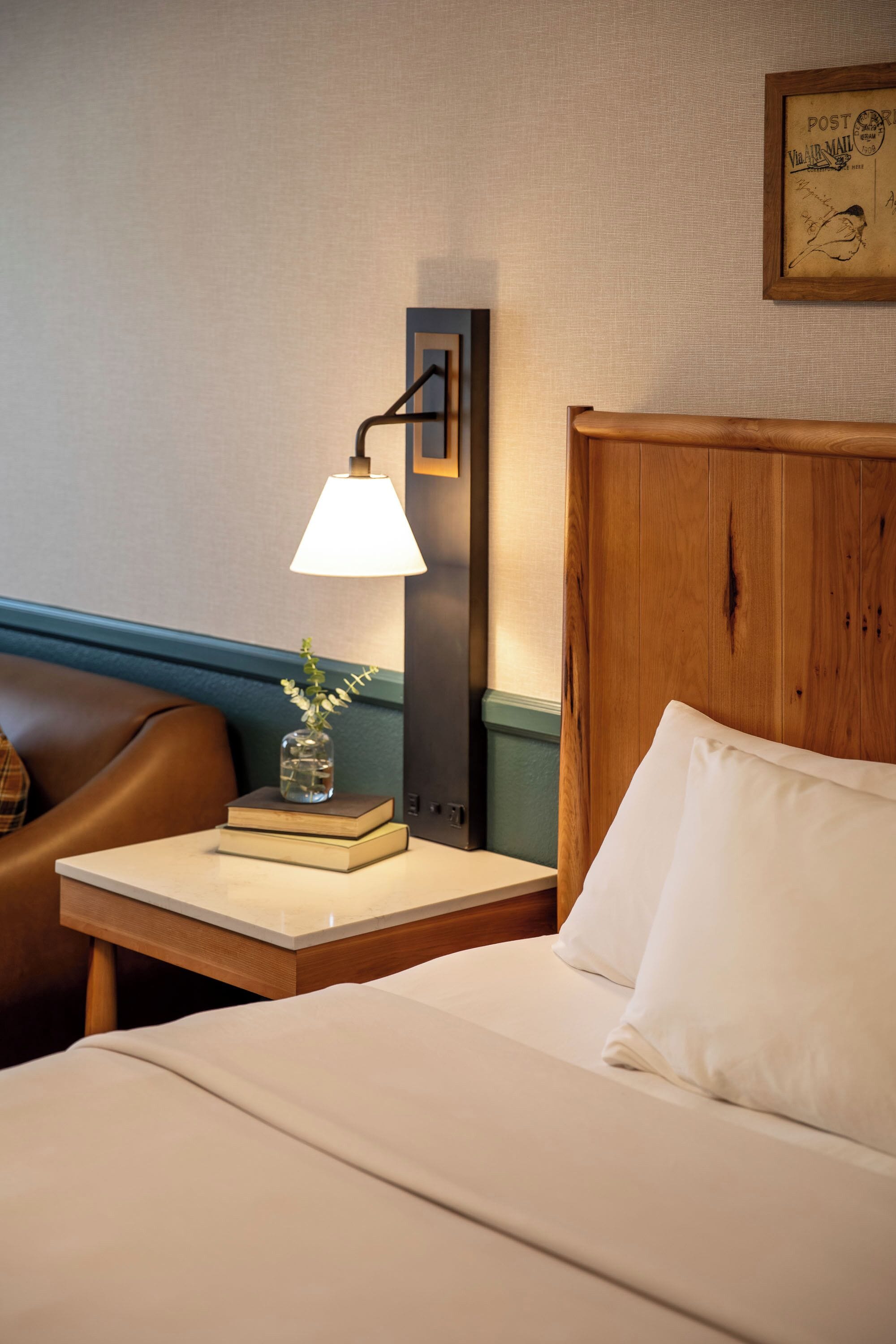 Une chambre d’hôtel avec un lit, une table de chevet avec une lampe, un vase de fleurs et un tableau encadré au mur.