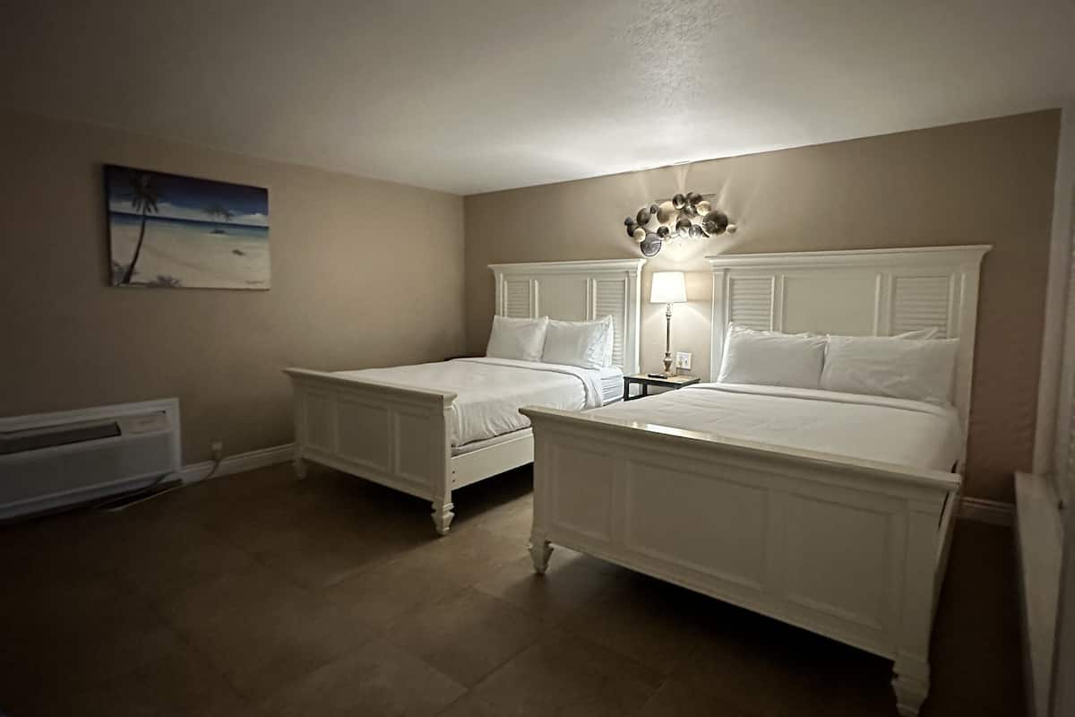 A bedroom with two white beds, a nightstand with a lamp, a painting of a beach scene, and a radiator on the wall.