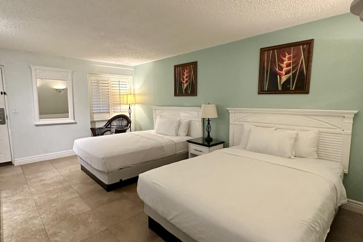 A hotel room with two beds, a window with blinds, a chair, a nightstand, and artwork on the walls.