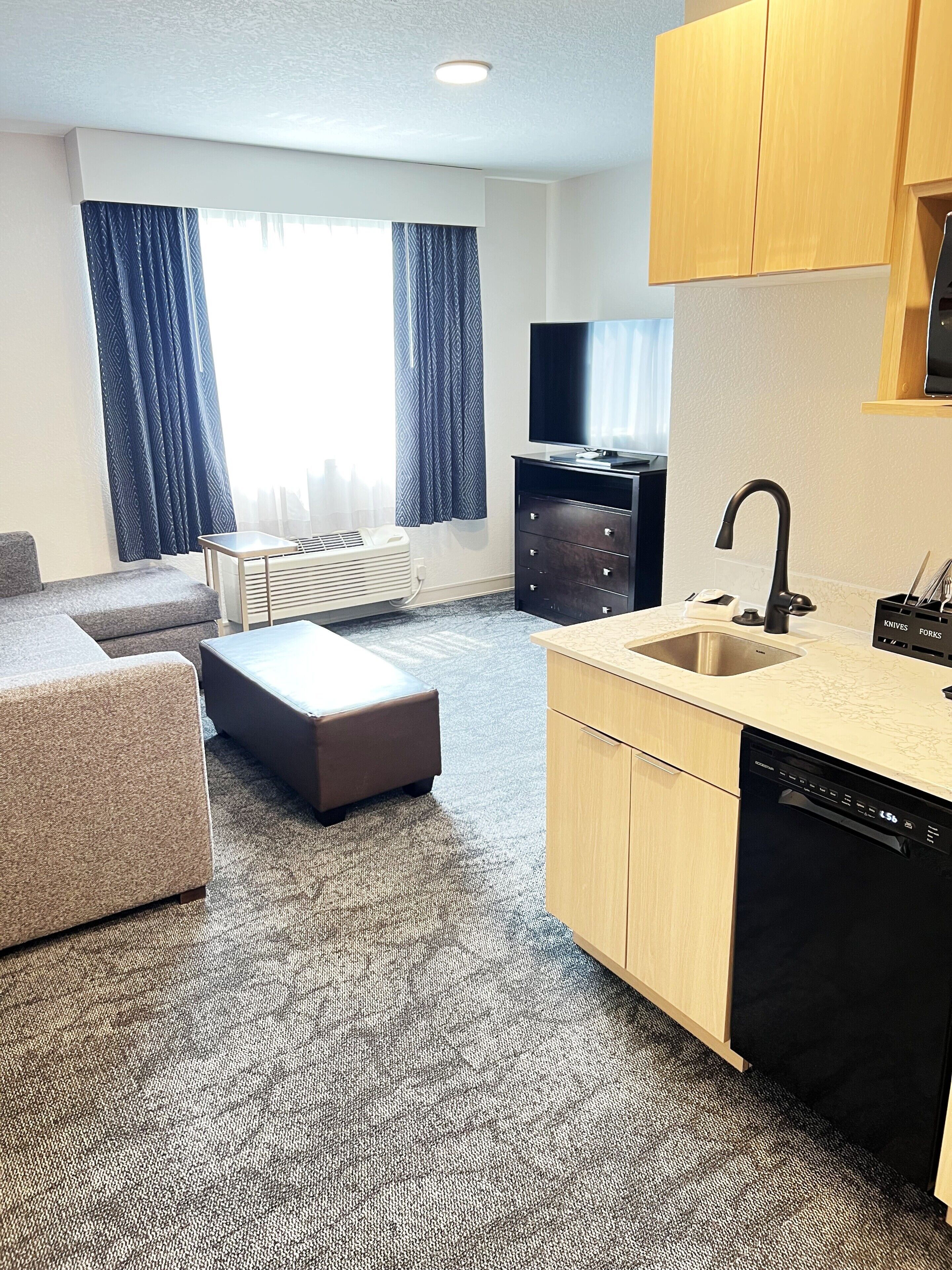 A modern hotel room with a kitchenette, a sofa, a flat-screen TV, and a large window with curtains.