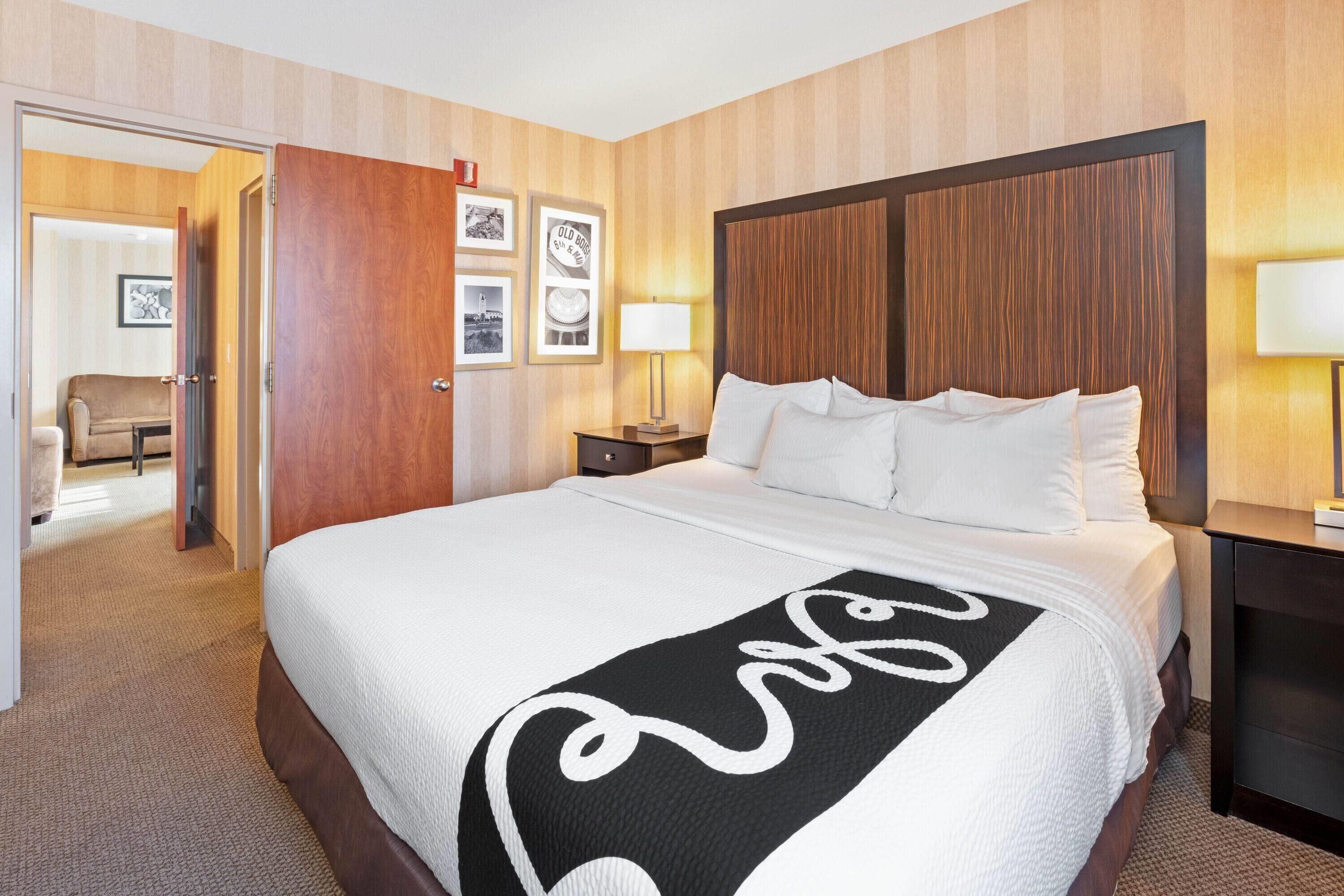 A hotel room with a large bed, wooden headboard, a bedside table with a lamp, and a door leading to a sitting area.