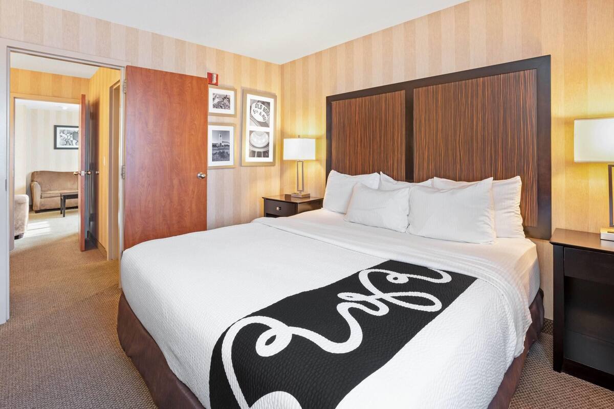 A hotel room with a large bed, wooden headboard, a bedside table with a lamp, and a door leading to a sitting area.