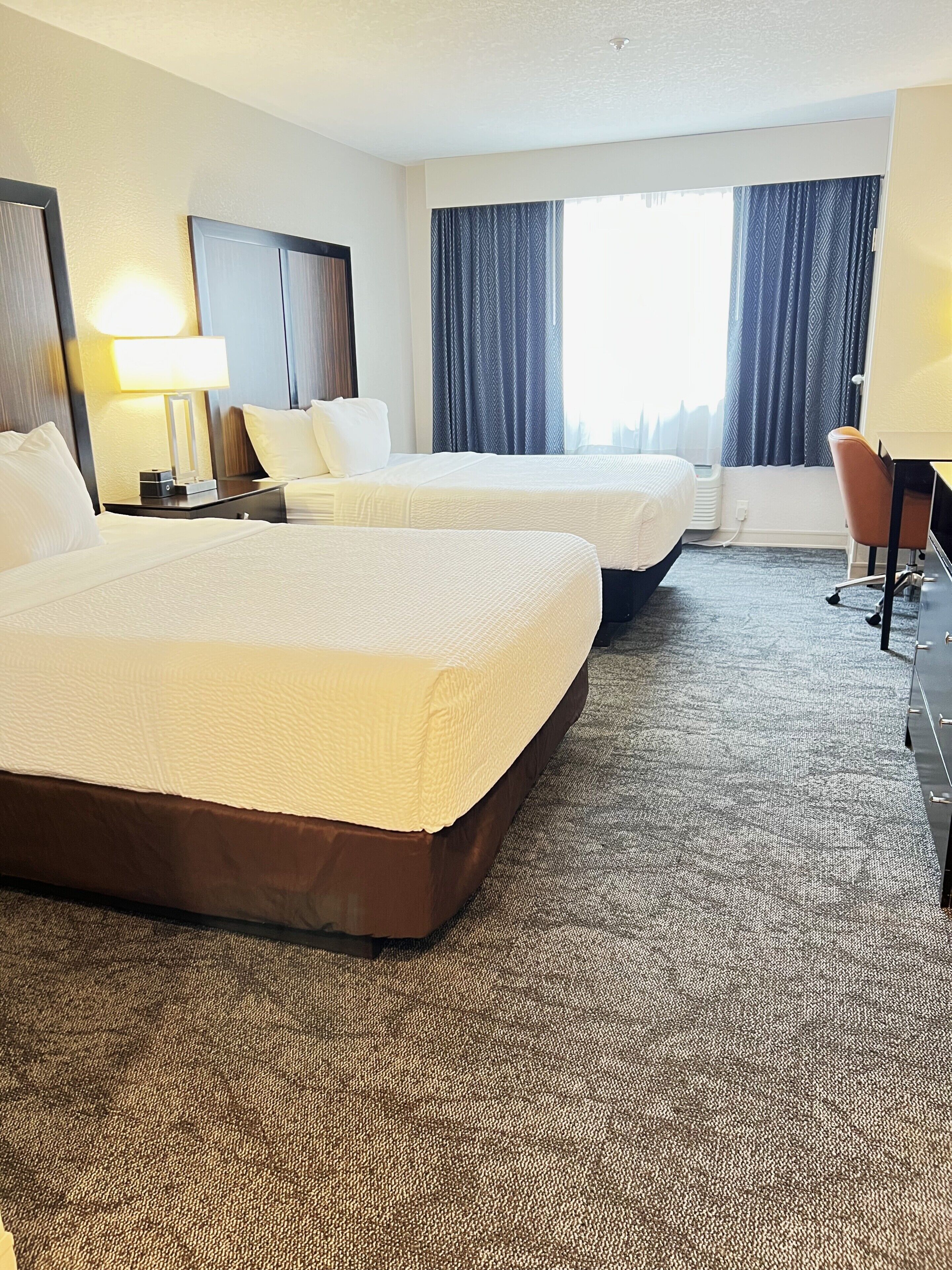A hotel room with two beds, a desk, a chair, and a window with curtains.