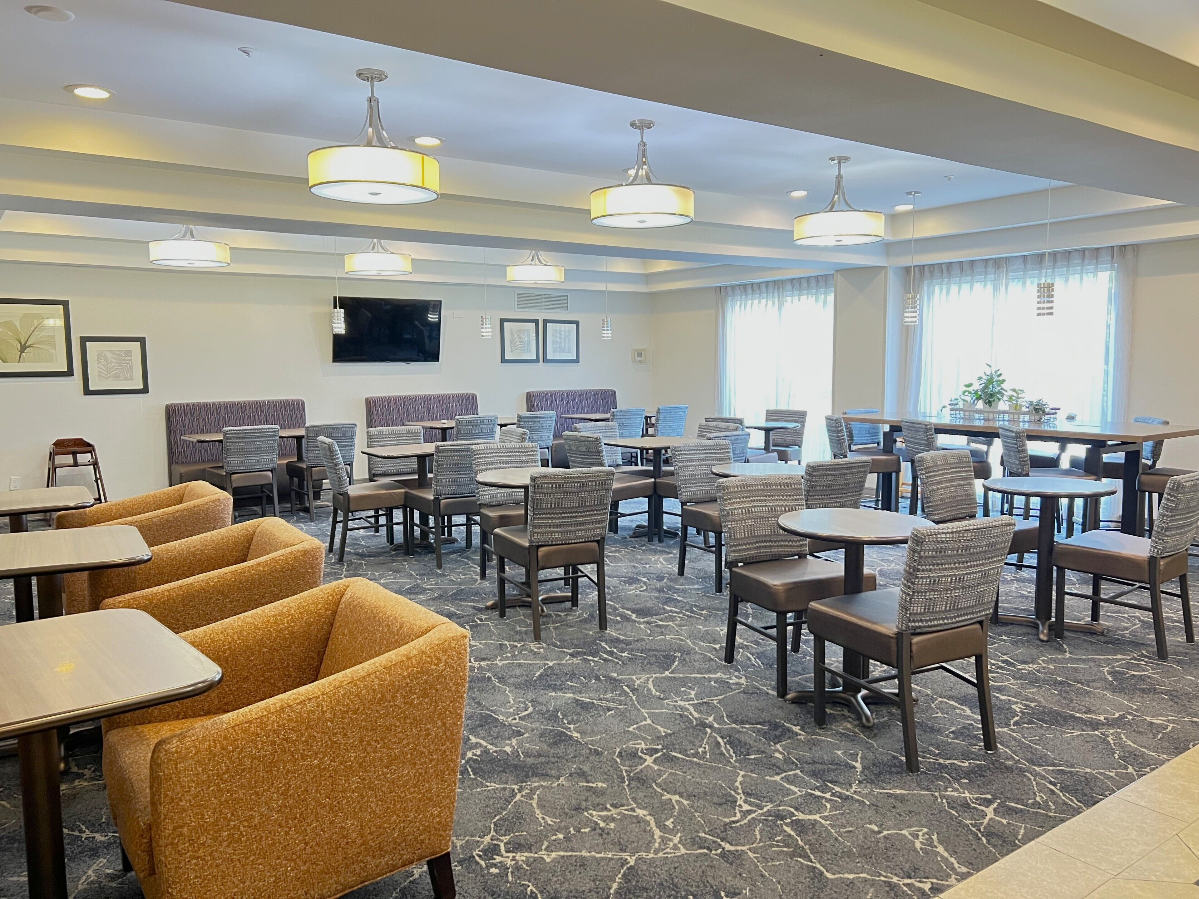 Une salle à manger d’hôtel avec de nombreuses tables et chaises, un téléviseur à écran plat fixé au mur et de grandes fenêtres laissant entrer la lumière naturelle.