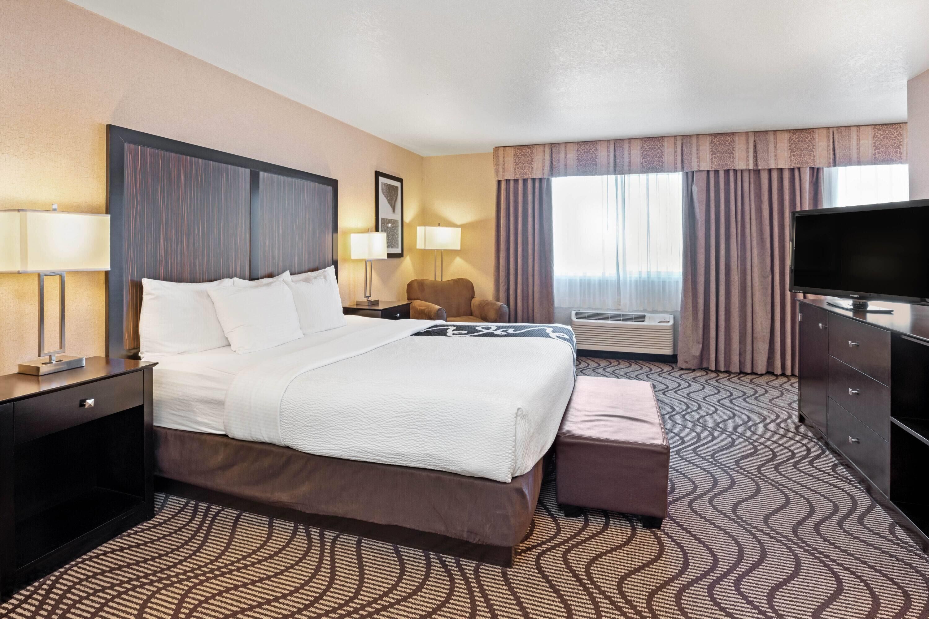 A hotel room with a bed, bedside tables, a TV, a chair, and a window with curtains.