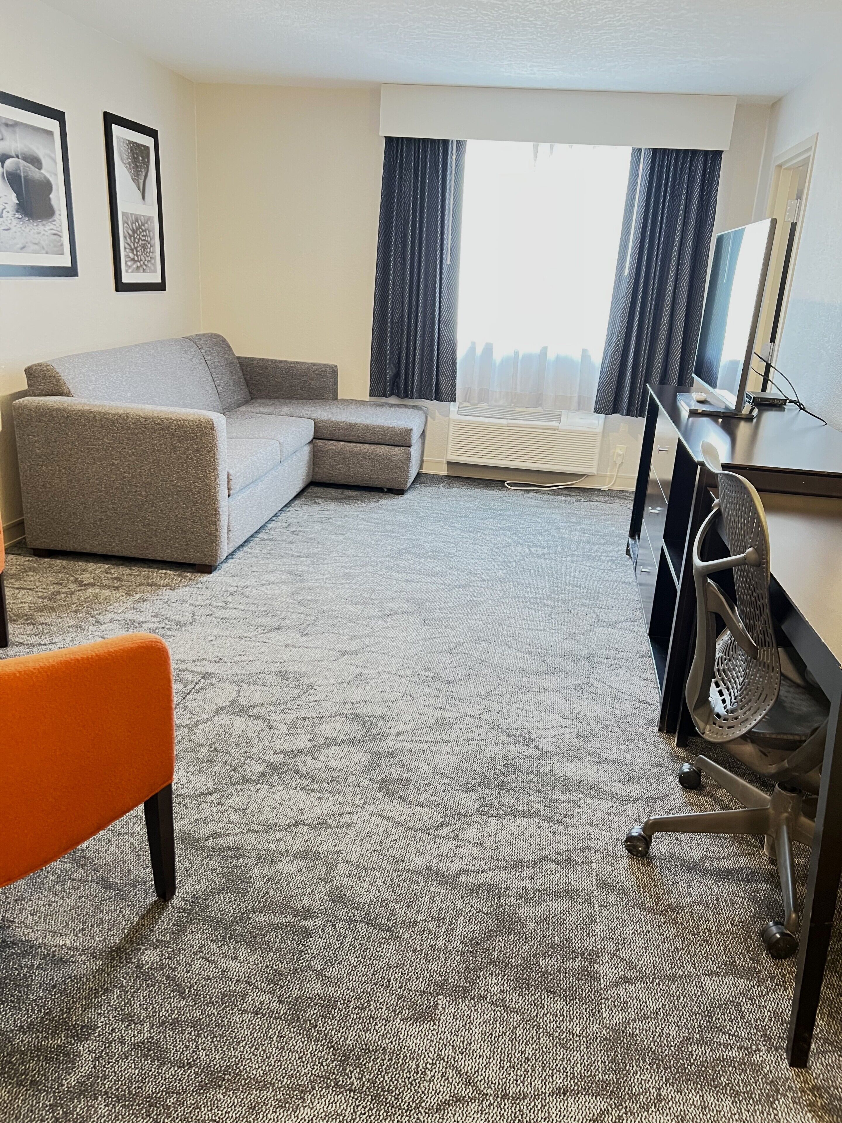 Ein Hotelzimmer mit grauem Sofa, einem orangefarbenen Sessel, einem Schreibtisch mit Computer und einem großen Fenster mit Vorhängen.