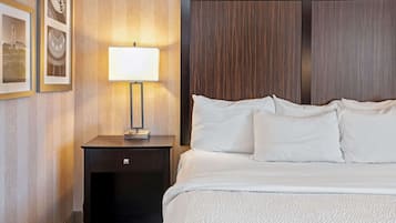 Un letto rifatto con lenzuola bianche e una testiera scura. Si nota un comodino con una lampada e una cornice con un'immagine.