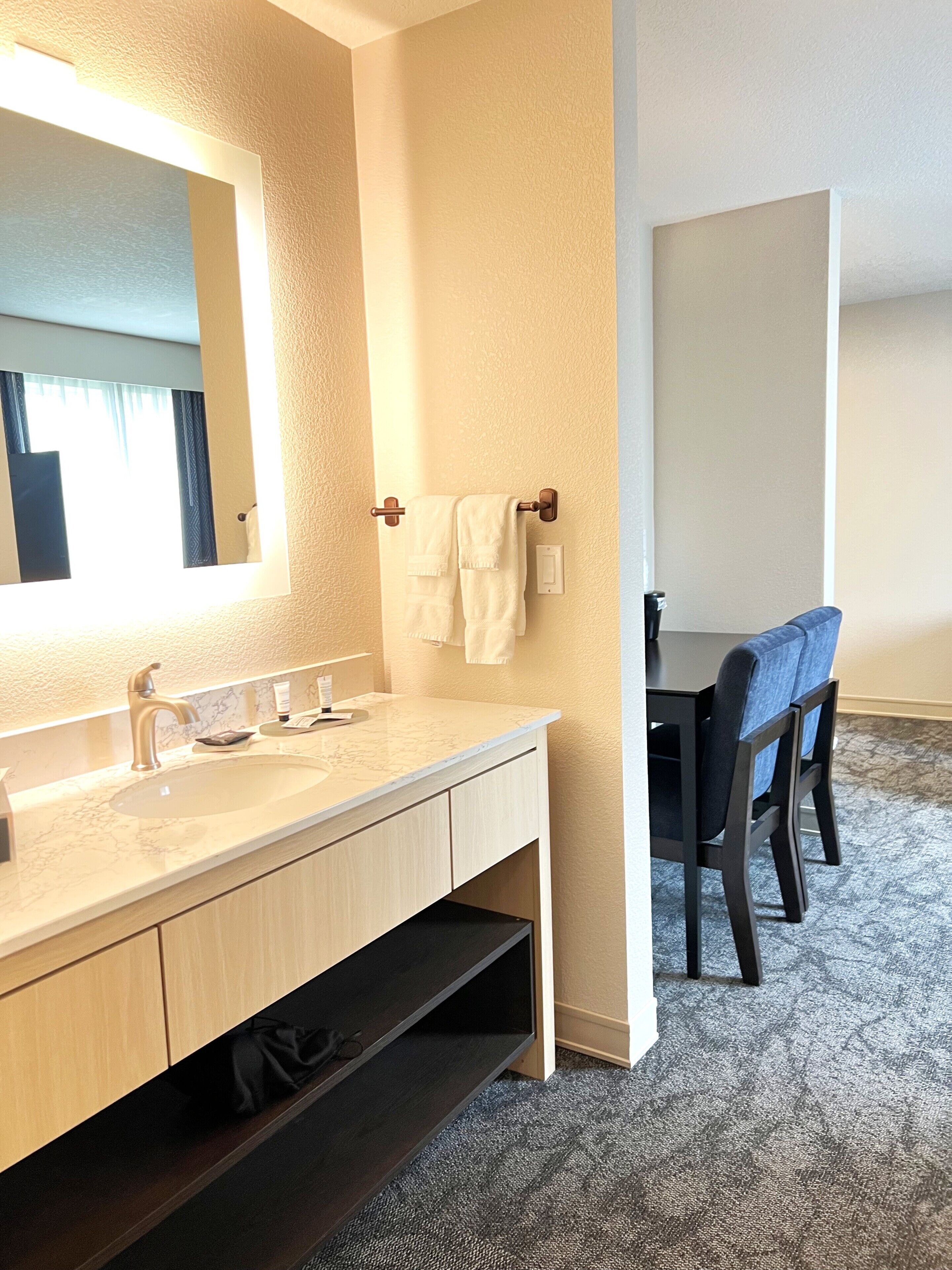 Un baño de hotel con lavamanos de mármol, un espejo grande, un perchero y un área de comedor con una mesa y sillas.