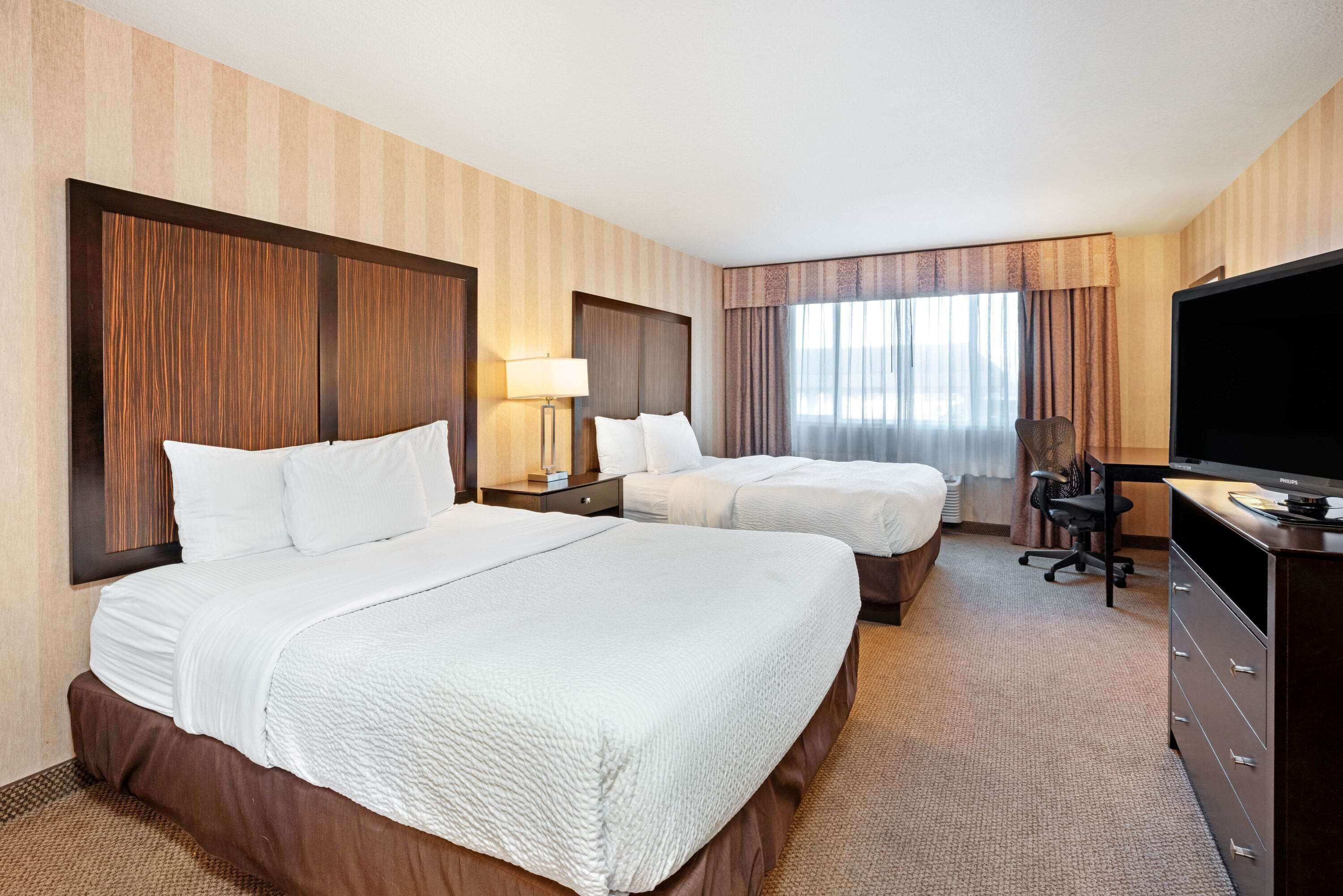 A hotel room with two beds, a TV, a desk, and a window with curtains.