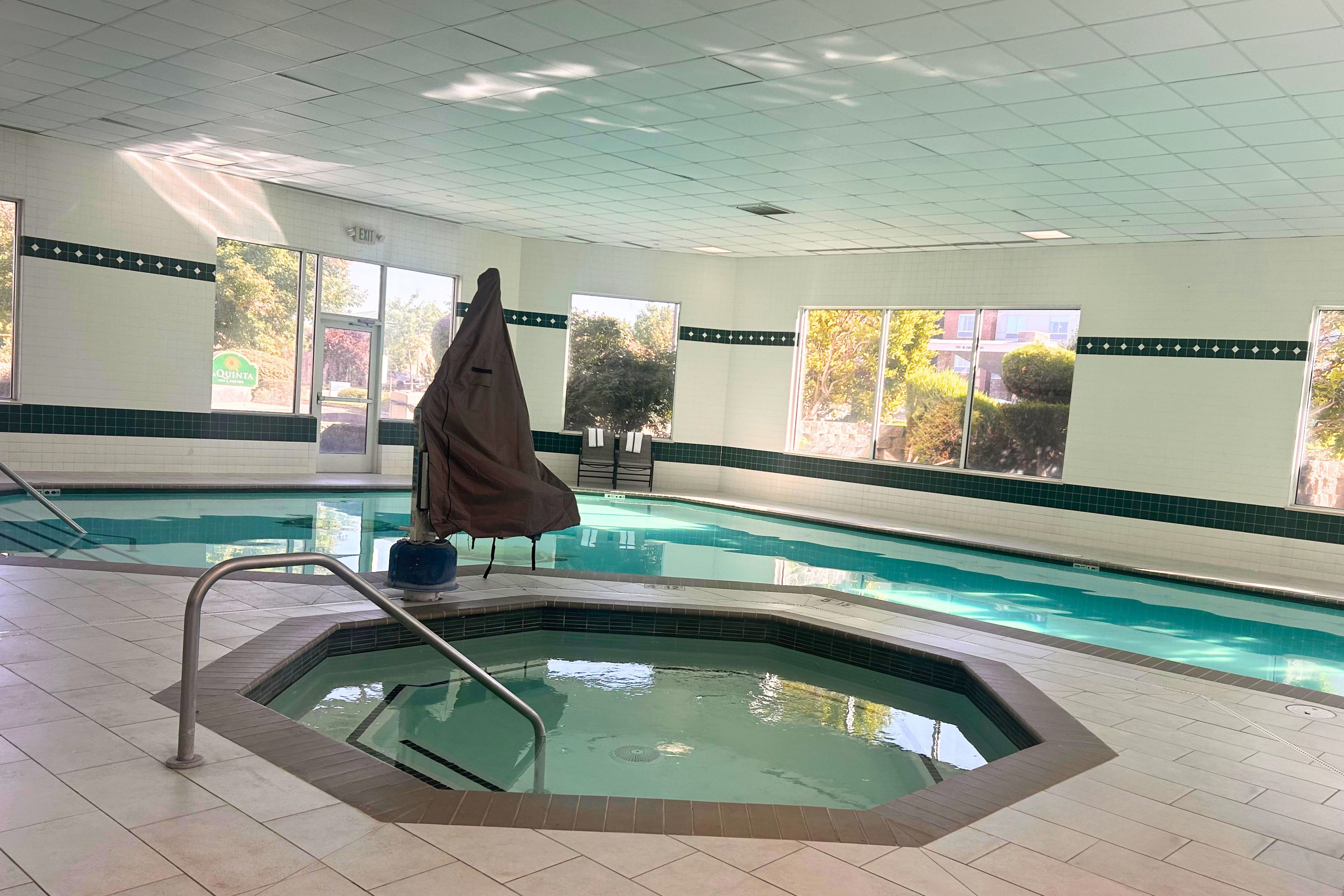 An indoor swimming pool with a hot tub, tiled walls, and windows with views of trees.