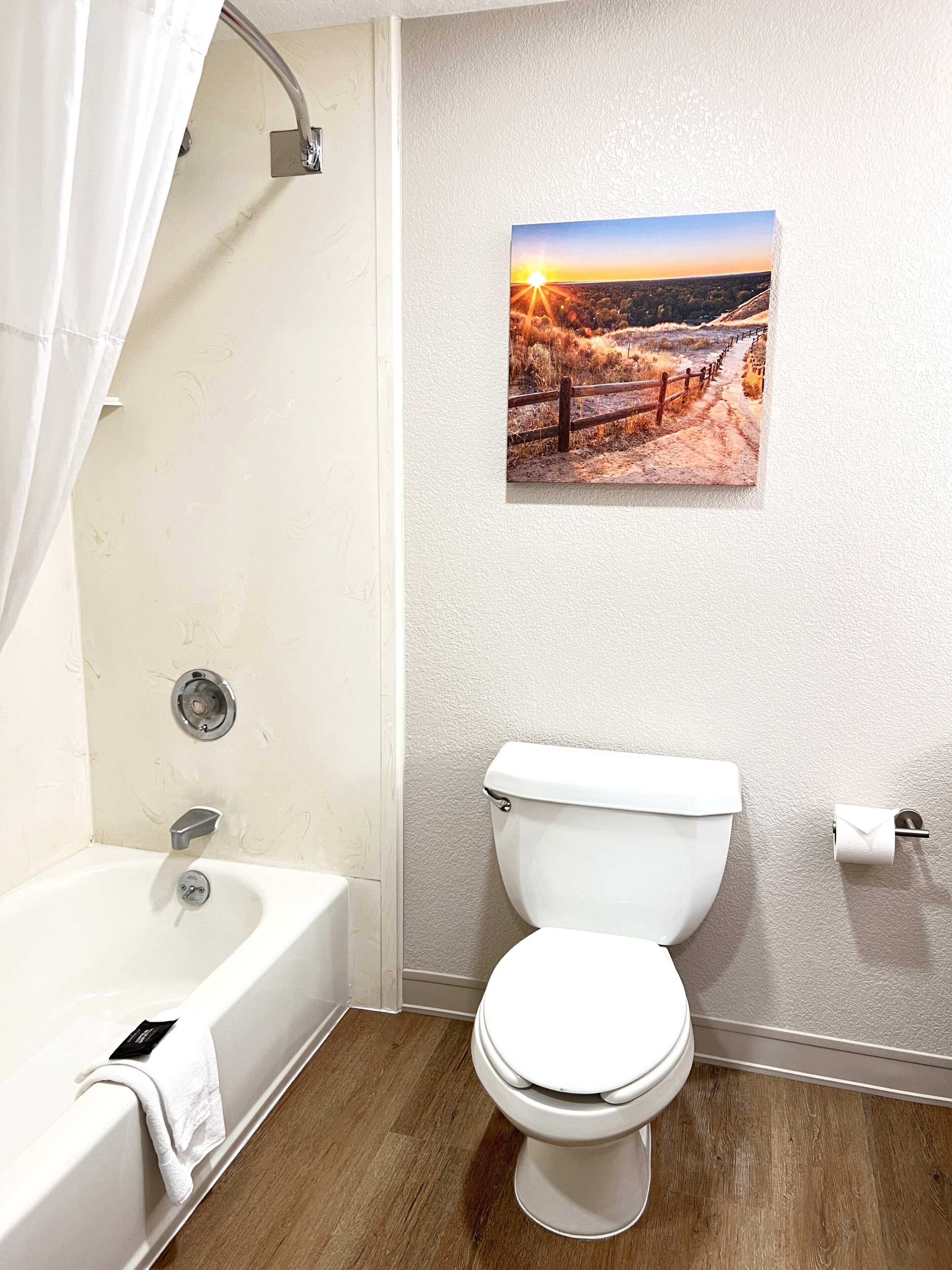 Een badkamer met een bad, toilet en een foto van een zonsonderganglandschap aan de muur.