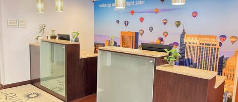 A hotel reception area with a modern design, featuring a reception desk, a digital screen, and a wall with a cityscape and hot air balloon decoration.