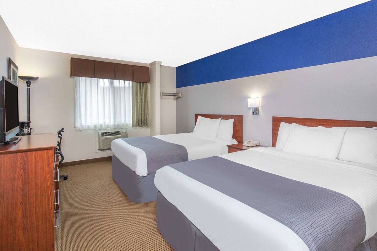 A hotel room with two beds, a desk with a TV, an air conditioning unit, and a window with curtains.