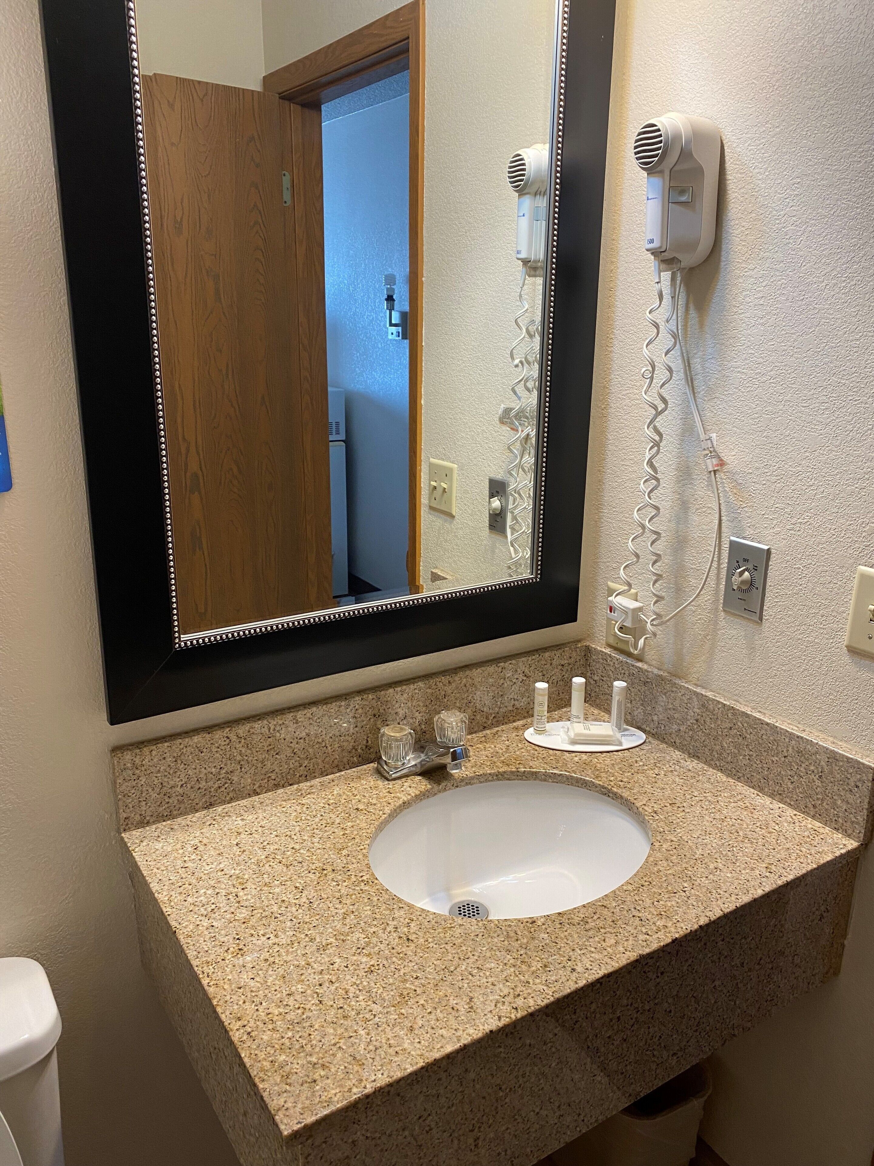 Une salle de bain dotée d’un lavabo avec un plan de travail en granit, d’un grand miroir avec un cadre décoratif, d’un sèche-cheveux et d’un porte-serviettes.