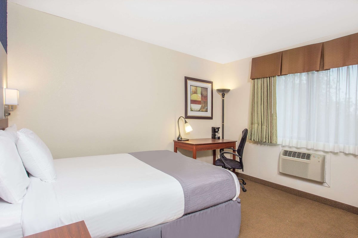 A hotel room with a large bed, a desk with a chair, a lamp, a framed picture on the wall, and a window with curtains.