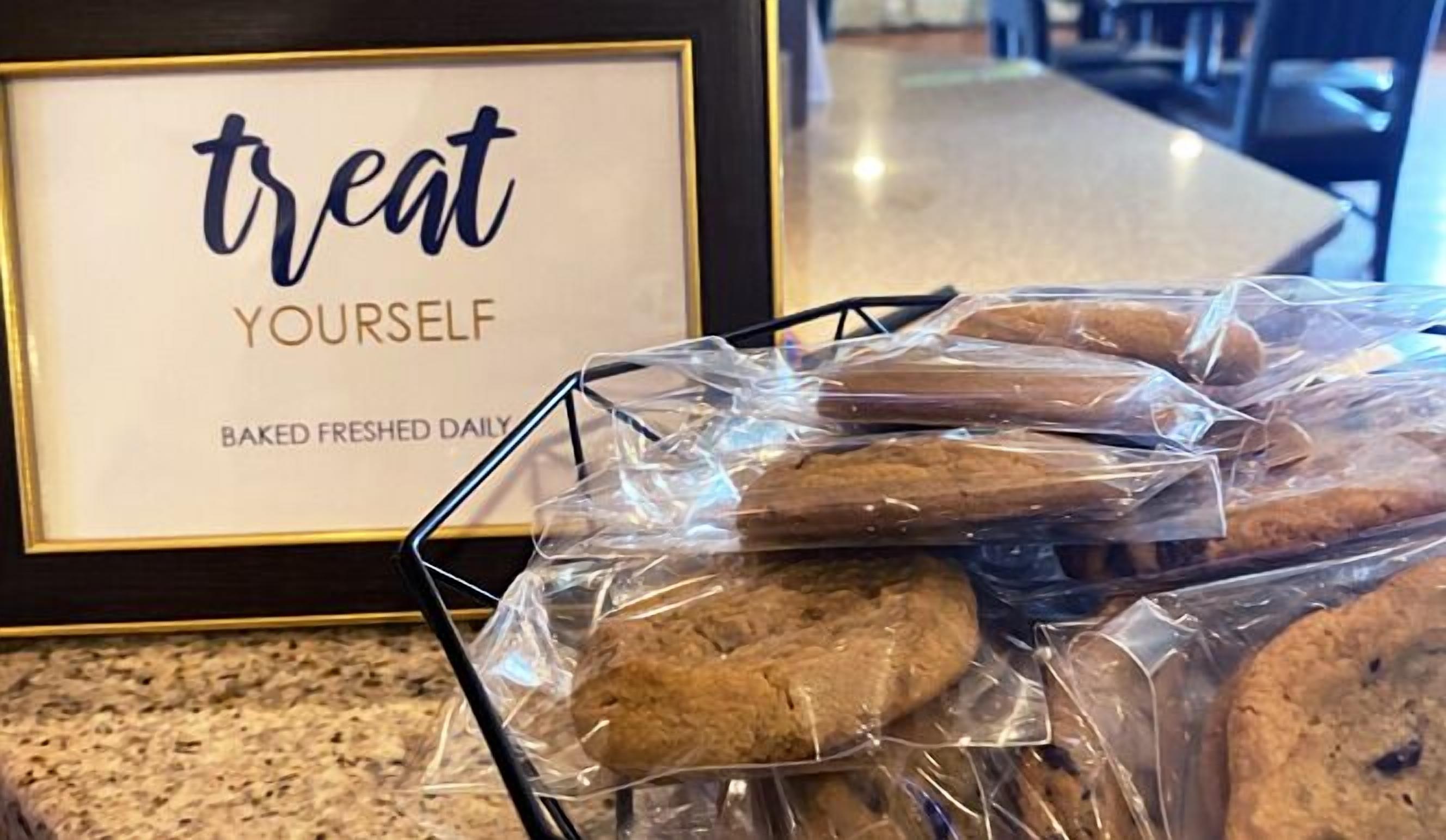 Ein gerahmtes Schild mit der Aufschrift "treat YOURSELF" und "BAKED FRESHEN DAILY" neben einem Metallkorb mit mehreren in Folie verpackten Keksen.