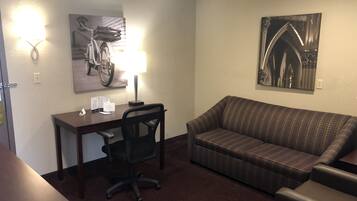 A hotel room with a desk, office chair, and a sofa. There are wall-mounted lights and two framed pictures.