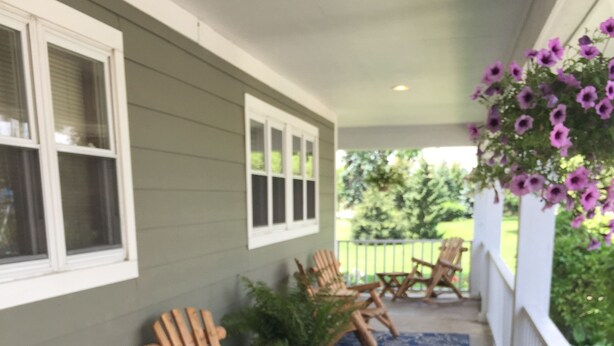 A covered porch with wooden Adirondack chairs, a patterned rug, and a flower basket.