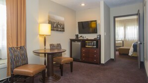 A hotel room with a dining table, two chairs, a microwave, a refrigerator, a television, and a bed.