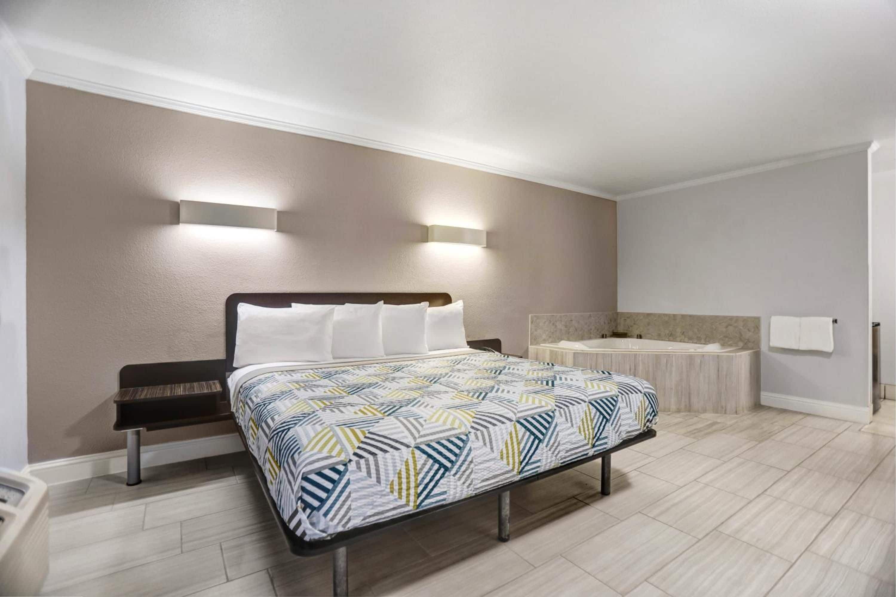 A hotel room with a bed, a nightstand, a wall-mounted light, a bathtub, and a towel rack.