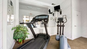 Une salle de sport équipée d’un tapis de course, d’un vélo elliptique et d’un vélo d’appartement. On y trouve une plante en pot et une porte sur laquelle est apposée une pancarte indiquant la sortie.