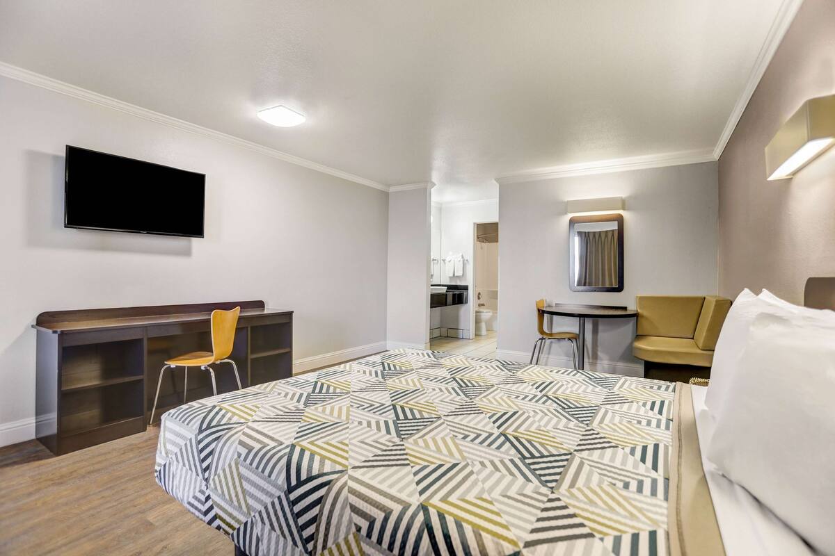 A hotel room with a bed, a desk, a chair, a TV, and a bathroom visible through an open door.