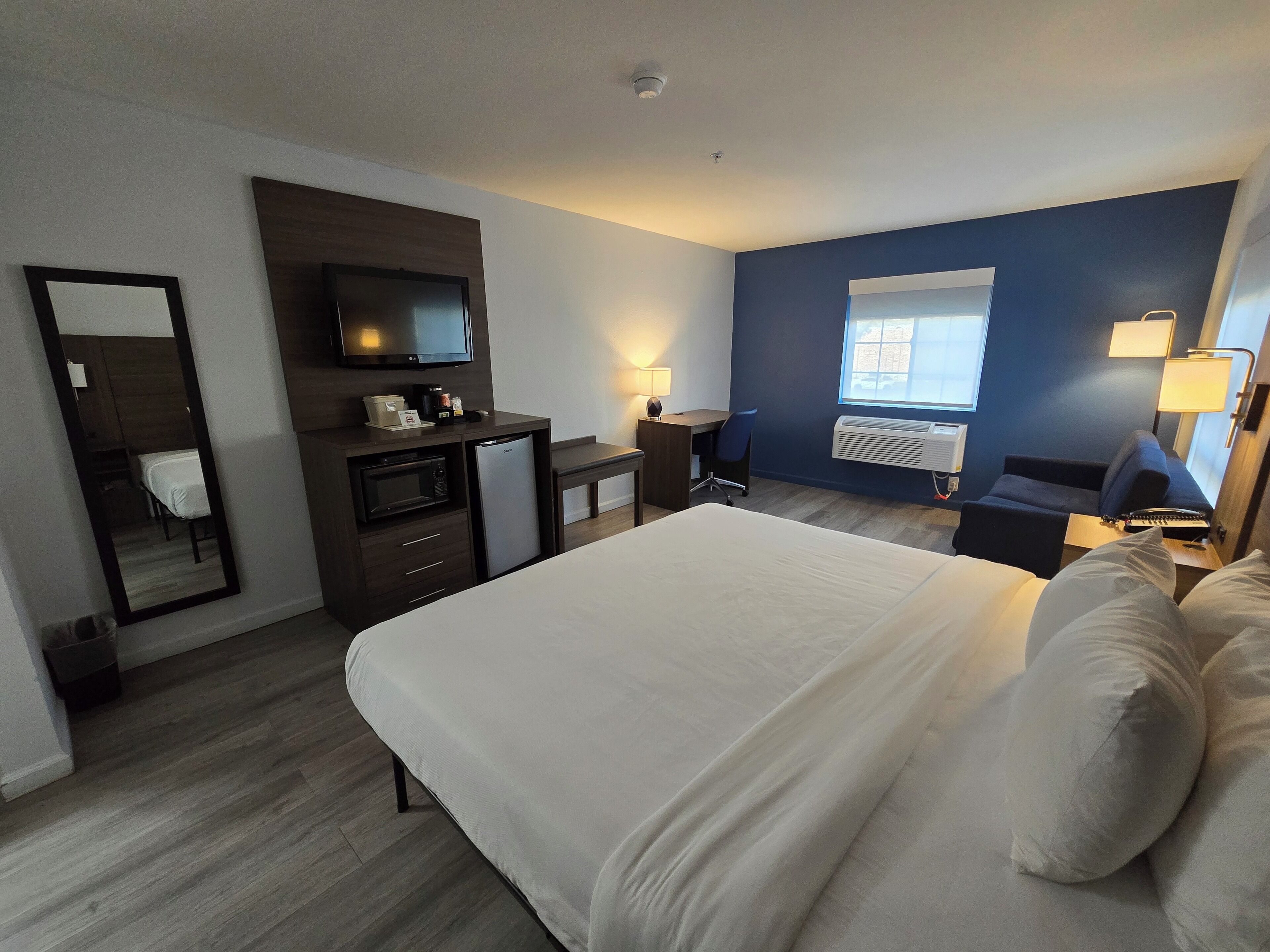 Una camera d'albergo con un letto, una scrivania, un televisore, uno specchio, una finestra e un condizionatore d'aria a muro.