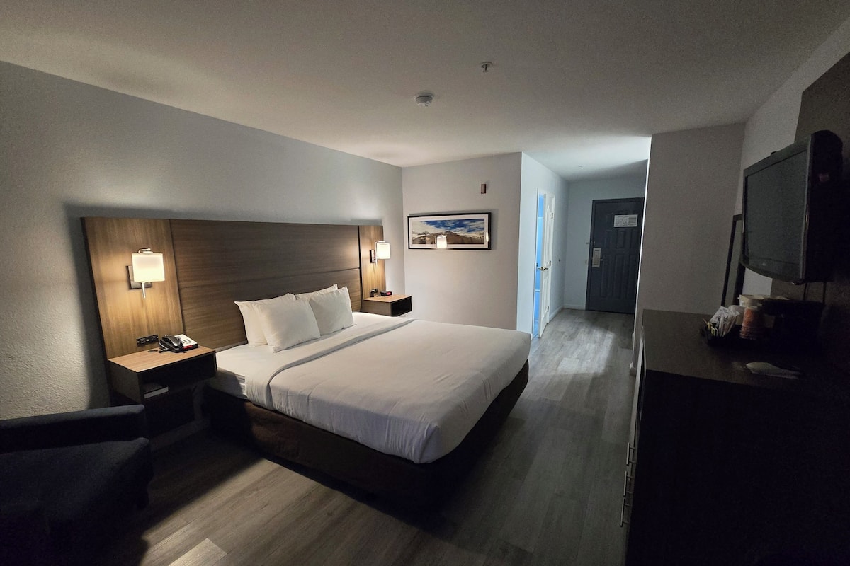 Una habitación de hotel con una cama grande, mesitas de noche, un escritorio con una silla, un televisor y una puerta que da a otra habitación.