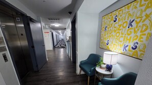 Un couloir d’hôtel avec un plancher en bois, une chaise bleue, une petite table avec une lampe et un mur orné d’une fresque représentant l’alphabet en jaune.