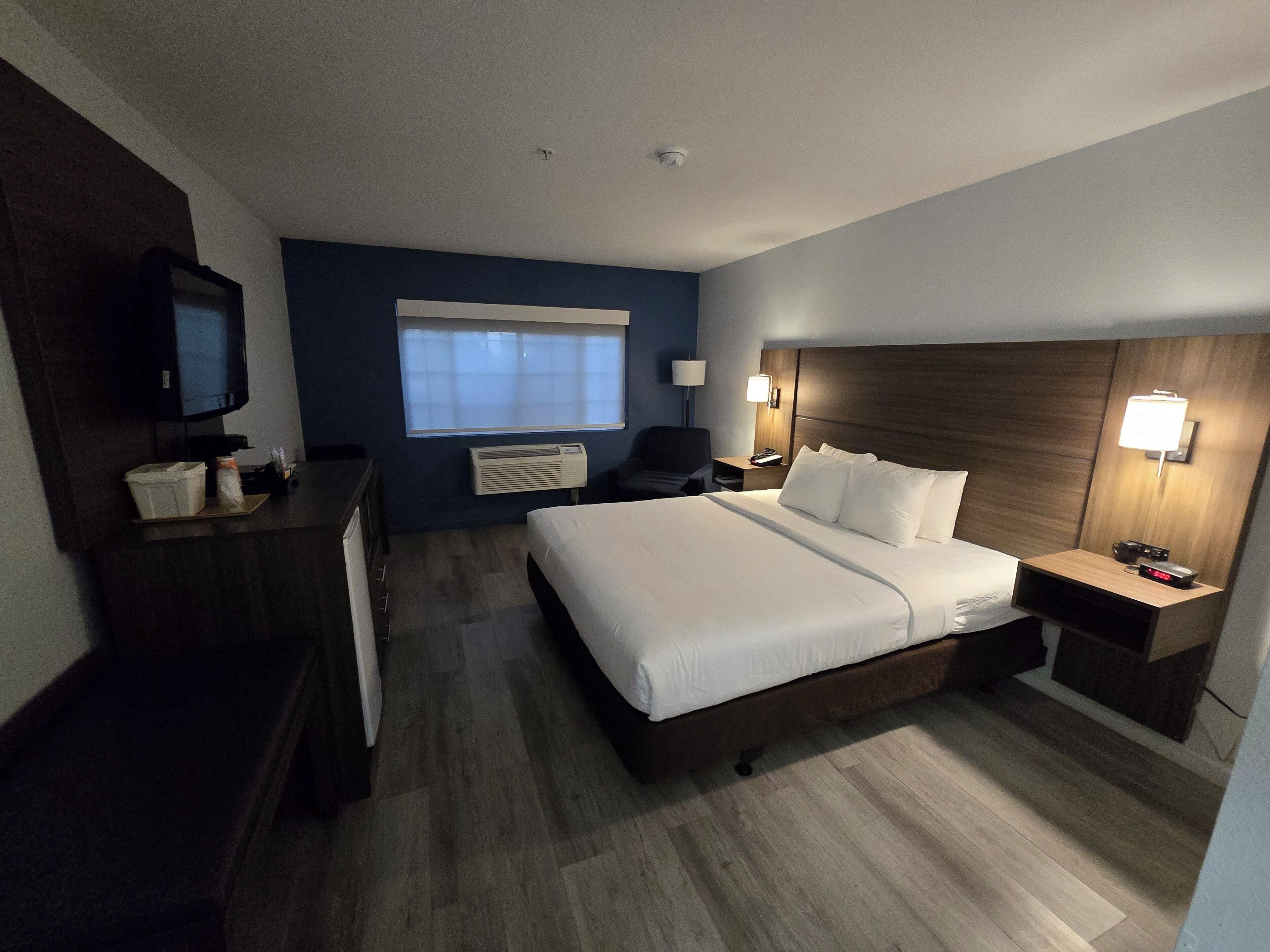 A hotel room with a bed, a desk, a chair, a TV, a lamp, and a nightstand with a clock.