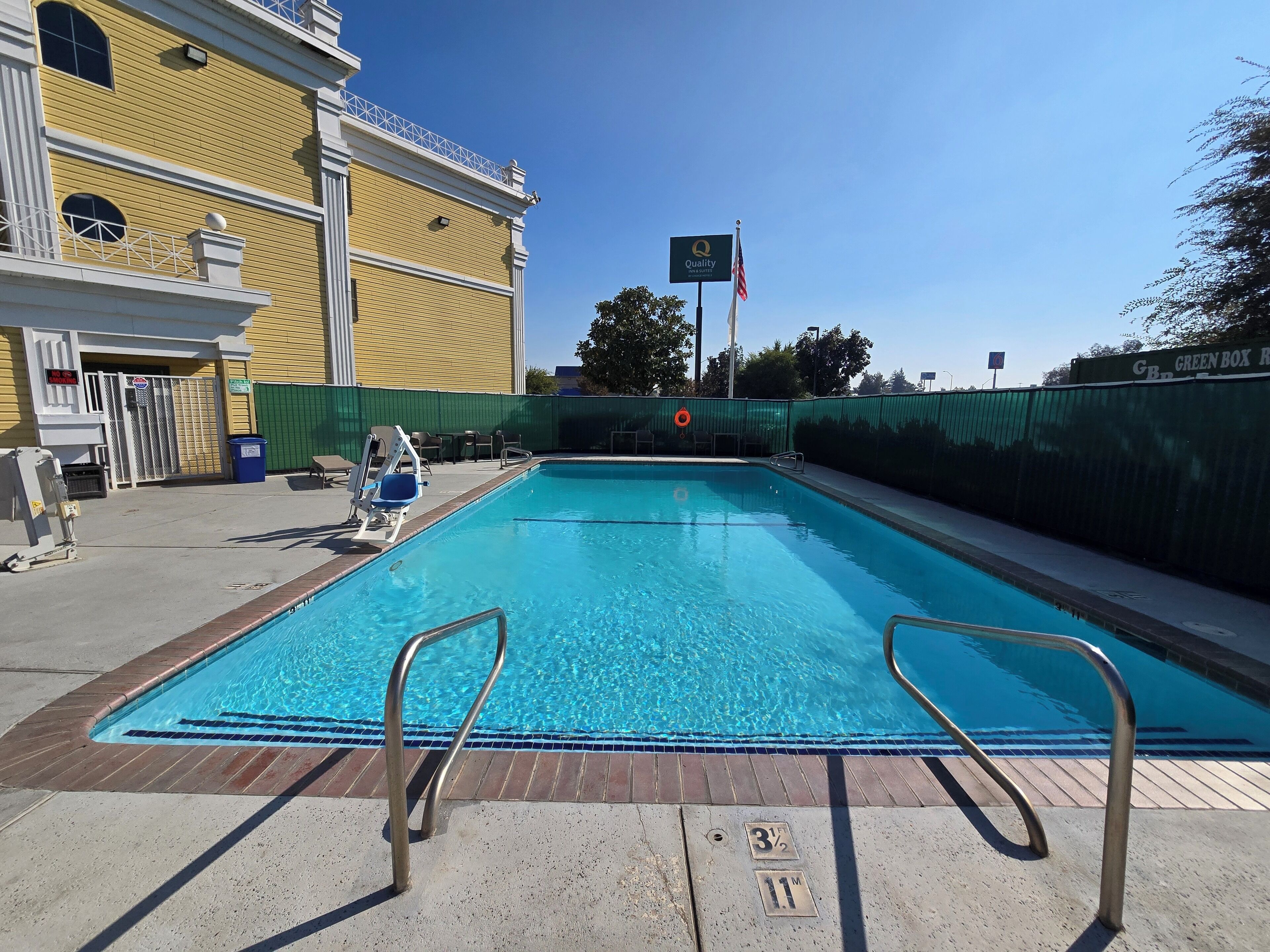 A swimming pool with metal handrails, a green fence, and a building with a yellow facade in the background.