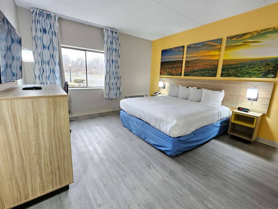 Et hotelværelse med seng, natbord, fjernsyn og et vindue med gardiner.